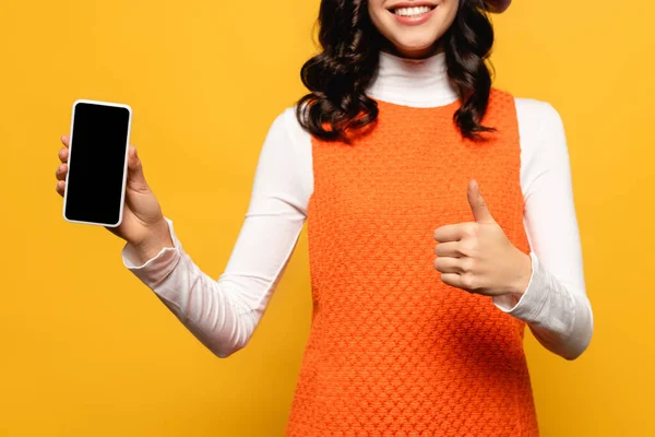 Vista recortada de mujer morena con el pulgar hacia arriba mostrando teléfono inteligente con pantalla en blanco aislado en amarillo - foto de stock