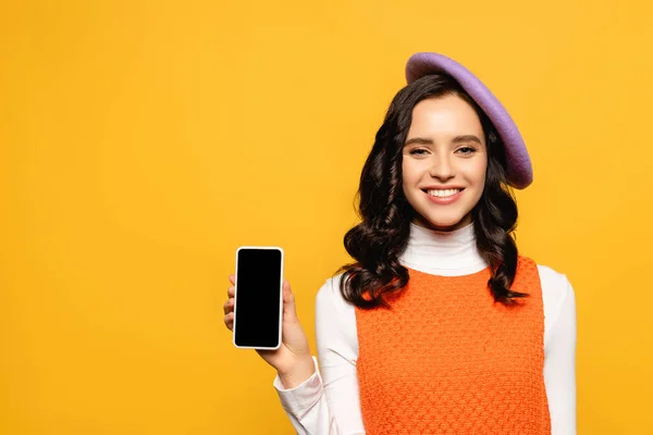 Glückliche brünette Frau mit Baskenmütze blickt in die Kamera, während sie ihr Smartphone isoliert auf gelb zeigt — Stockfoto