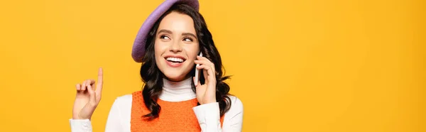 Happy brunette woman in beret with idea gesture talking on smartphone while looking away isolated on yellow, banner — Stock Photo