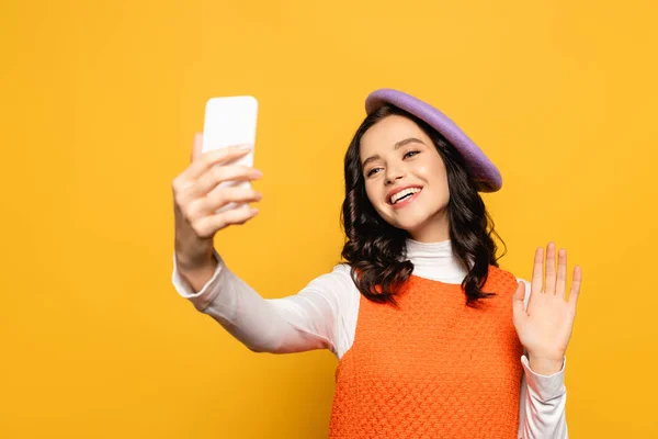 Fröhliche brünette Frau mit winkender Hand, Baskenmütze und Selfie auf gelb — Stockfoto