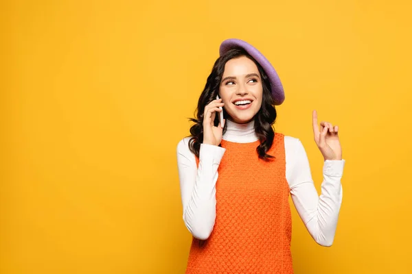 Mujer morena alegre en boina con gesto de idea hablando en el teléfono inteligente mientras mira hacia otro lado aislado en amarillo - foto de stock