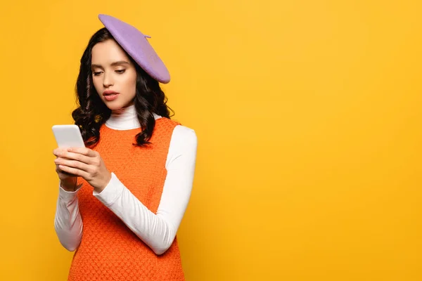 Lockige brünette Frau in Baskenmütze SMS auf Smartphone isoliert auf gelb — Stockfoto