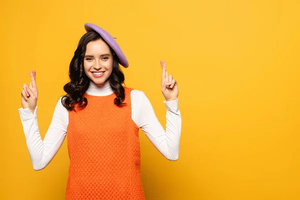 Happy brunette woman in beret with crossed fingers looking at camera isolated on yellow — Stock Photo