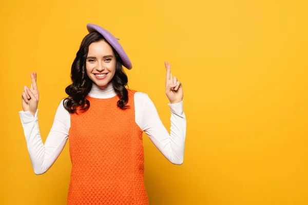 Mujer morena feliz en boina con los dedos cruzados mirando a la cámara aislada en amarillo - foto de stock