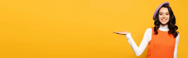 Щаслива брюнетка в Береті дивиться на камеру, вказуючи рукою ізольовано на жовтому, банер — стокове фото