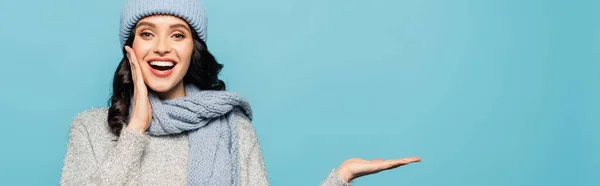 Glückliche brünette Frau mit offenem Mund im Winteroutfit und mit der Hand isoliert auf blau zeigend, Banner — Stockfoto