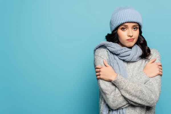Sad brunette woman in hat and scarf hugging herself while looking at camera isolated on blue — Stock Photo