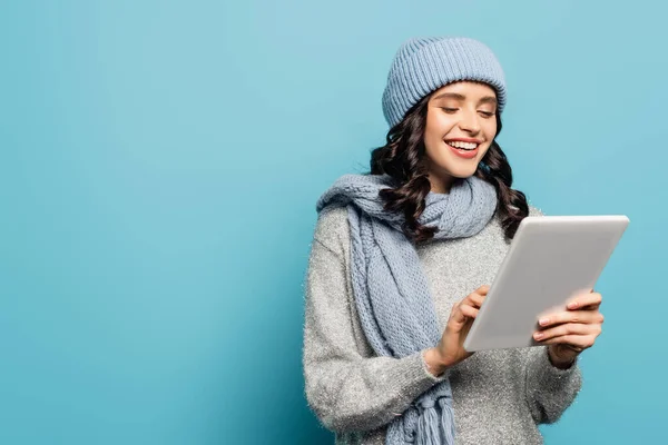 Mulher morena alegre em chapéu e cachecol usando tablet digital isolado em azul — Fotografia de Stock