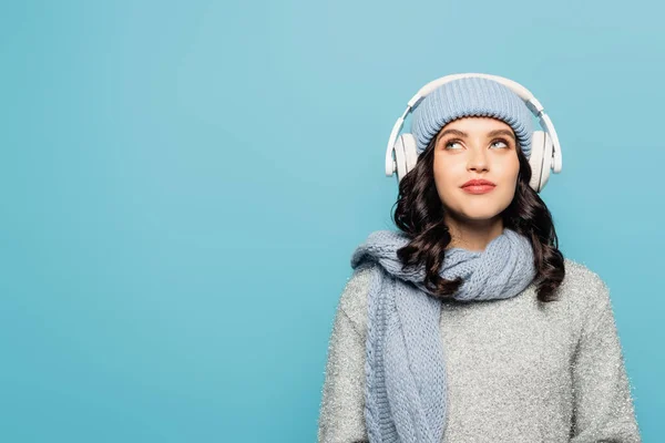 Verträumte Frau im winterlichen Outfit mit Kopfhörern denkt nach, während sie isoliert auf blau blickt — Stockfoto