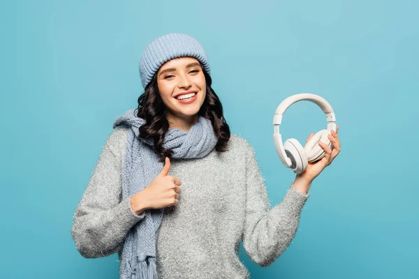 Mujer morena feliz en traje de invierno sosteniendo los auriculares y mostrando el pulgar hacia arriba aislado en azul - foto de stock