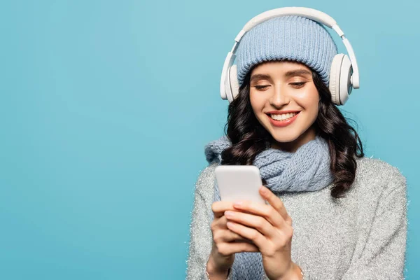 Mujer alegre en traje de invierno con auriculares mensajes de texto en el teléfono inteligente aislado en azul - foto de stock