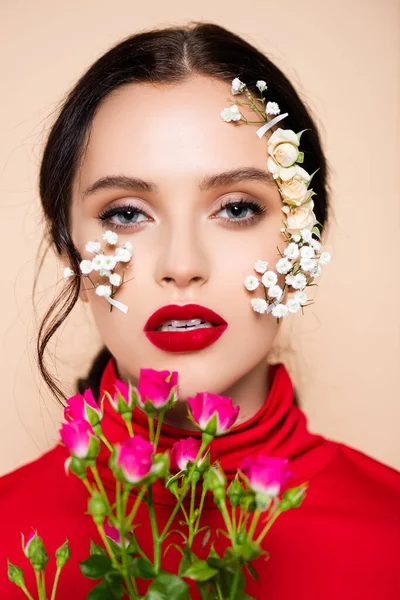 Mujer joven con labios rojos y flores en la cara mirando a la cámara aislada en rosa - foto de stock