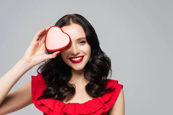 Fröhliche Frau mit roten Lippen, die Augen bedecken, während sie herzförmige Geschenkschachtel isoliert auf grau hält — Stockfoto