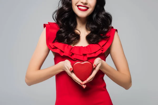 Vista parcial de mujer alegre con labios rojos sosteniendo caja de regalo en forma de corazón aislado en gris - foto de stock