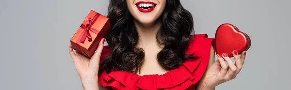 Cropped view of cheerful woman with red lips holding heart-shaped gift box and wrapped present with bow isolated on grey, banner - foto de stock