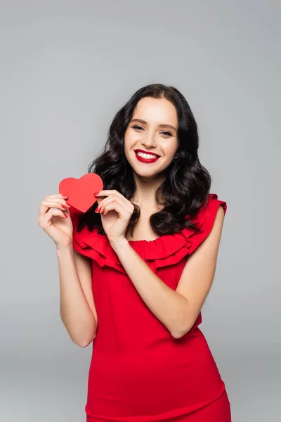 Alegre joven en vestido sosteniendo corazón de papel rojo aislado en gris - foto de stock