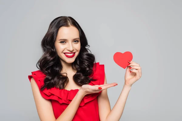 Усміхнена молода жінка вказує рукою на червоний папір серце ізольовано на сірому — стокове фото