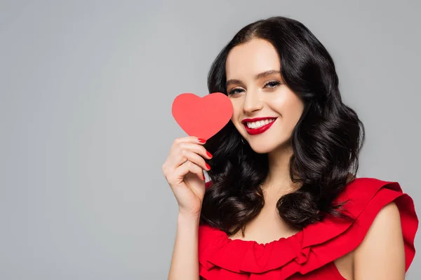 Усміхнена молода жінка тримає червоне паперове серце ізольовано на сірому — стокове фото