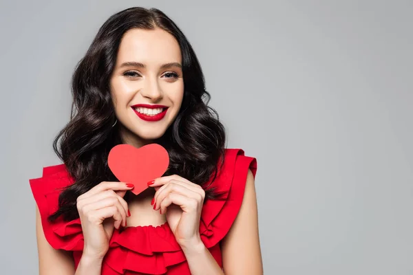 Sonriente morena mujer sosteniendo rojo papel corazón aislado en gris - foto de stock