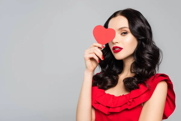 Joven morena mujer cubriendo ojo rojo papel corazón aislado en gris - foto de stock