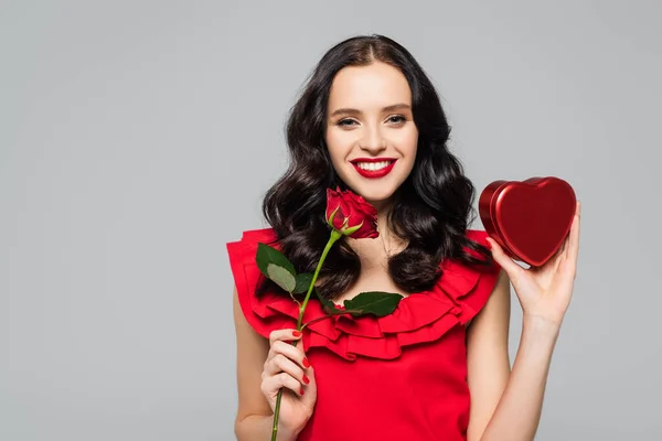 Gioiosa giovane donna che tiene rosa rossa e scatola a forma di cuore isolato su grigio — Foto stock