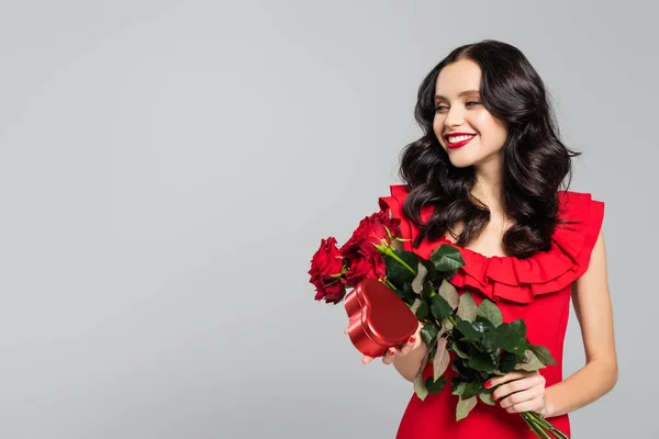 Heureuse jeune femme tenant bouquet de roses rouges et boîte en forme de coeur isolé sur gris — Photo de stock