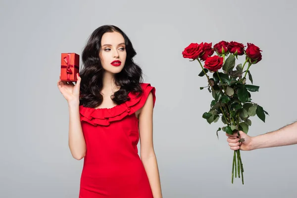 Man giving roses to brunette woman in red dress holding wrapped gift box isolated on grey — Stock Photo