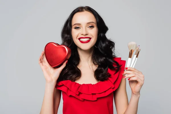 Веселая женщина с красными губами, держащая подарочную коробку в форме сердца и косметические щетки, изолированные на сером — стоковое фото