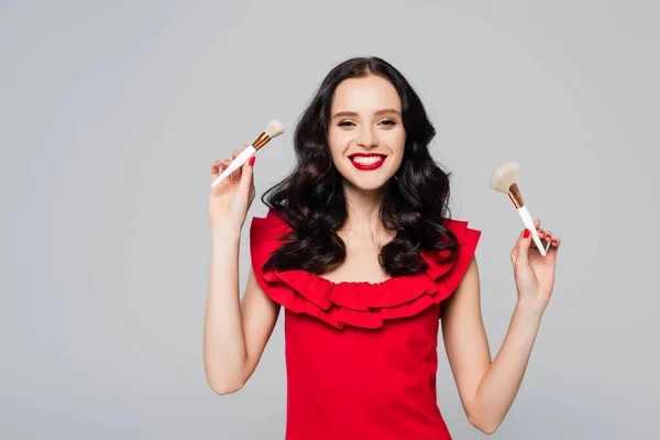 Cheerful woman with red lips holding cosmetic brushes isolated on grey — Stock Photo