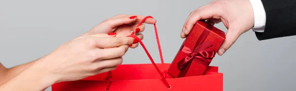 Vue recadrée de l'homme mettant boîte cadeau dans un sac en papier dans les mains de la femme avec des lèvres rouges isolées sur gris, bannière — Photo de stock