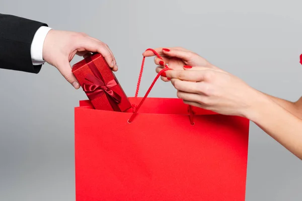 Cropped view of man putting gift box in paper bag in hands of woman with red lips isolated on grey — Stock Photo