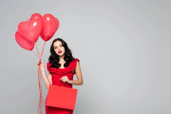 Mulher morena segurando balões em forma de coração vermelho e saco de compras isolado em cinza — Fotografia de Stock