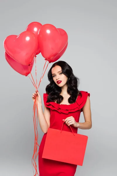 Mujer joven sosteniendo globos rojos en forma de corazón y bolsa aislada en gris - foto de stock