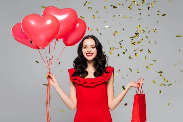 Mujer feliz sosteniendo globos rojos en forma de corazón y bolsa de compras cerca de caer confeti en gris - foto de stock