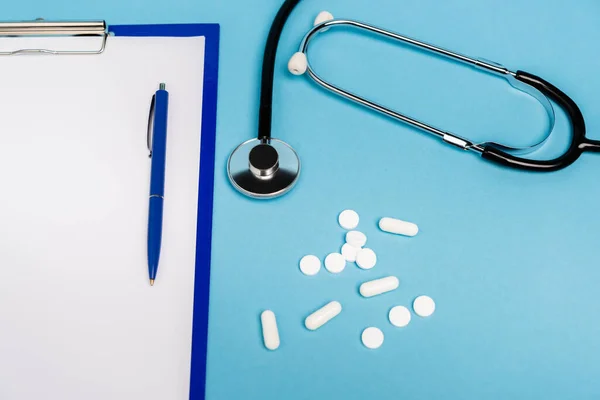 Vista ad alto angolo degli appunti con penna, pillole e stetoscopio su sfondo blu — Foto stock