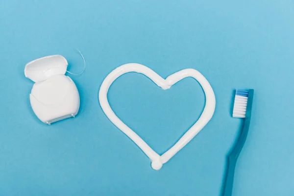 Vista dall'alto del segno del cuore dal dentifricio vicino allo spazzolino e al filo interdentale su sfondo blu — Foto stock