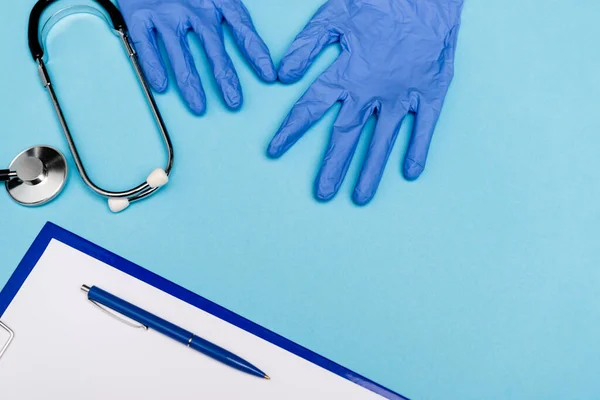 Vista superior de guantes de látex cerca del estetoscopio y portapapeles con pluma sobre fondo azul - foto de stock