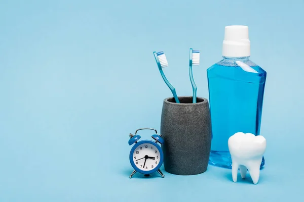 Reloj pequeño cerca de cepillos de dientes, enjuague bucal y modelo de dientes sobre fondo azul - foto de stock