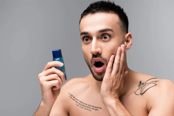 Schockierter hispanischer Mann berührt Gesicht, während er Flasche Rasierwasser isoliert auf grau hält — Stockfoto