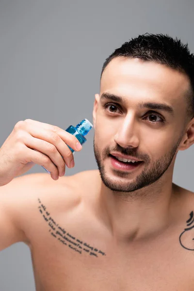 Sonriente, hombre hispano tatuado sosteniendo un frasco de agua de colonia aislado en gris - foto de stock
