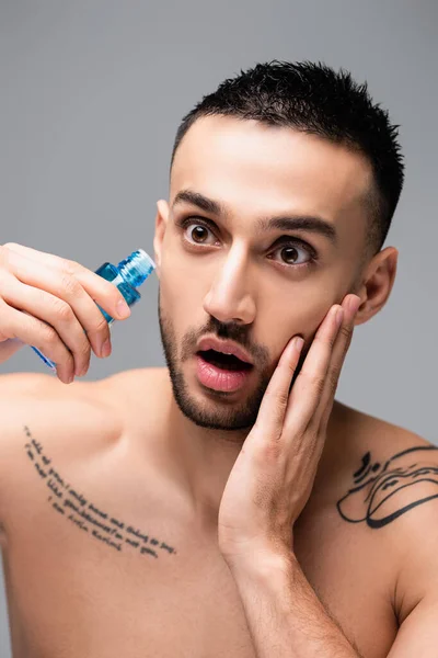 Asombrado hombre hispano tocando la cara mientras se aplica loción aftershave aislado en gris — Stock Photo
