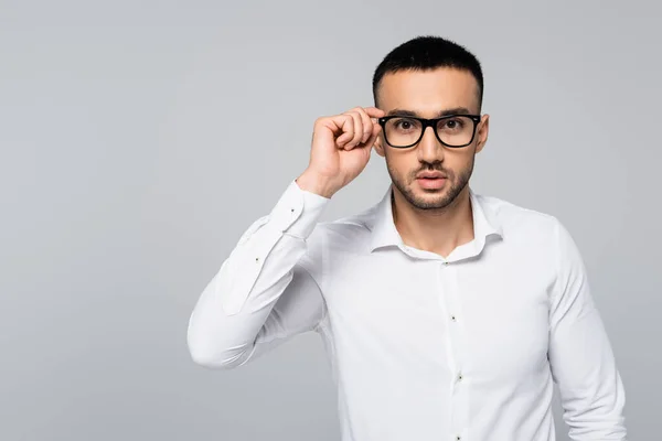 Junger hispanischer Manager in weißem Hemd, der seine Brille justiert, während er isoliert auf graue Kamera blickt — Stockfoto