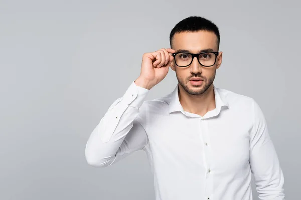 Joven gerente hispano en camisa blanca ajustando gafas mientras mira la cámara aislada en gris - foto de stock