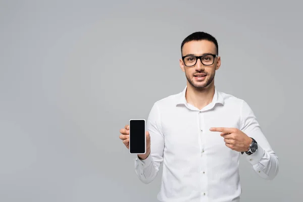 Joven empresario hispano con gafas apuntando con el dedo al smartphone con pantalla en blanco aislada en gris - foto de stock