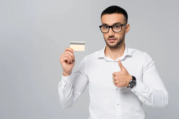 Jovem empresário hispânico mostrando polegar para cima enquanto segurando cartão de crédito isolado em cinza — Fotografia de Stock
