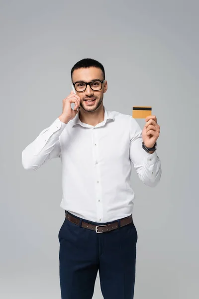 Homem de negócios hispânico positivo falando no celular enquanto mostra cartão de crédito isolado no cinza — Fotografia de Stock