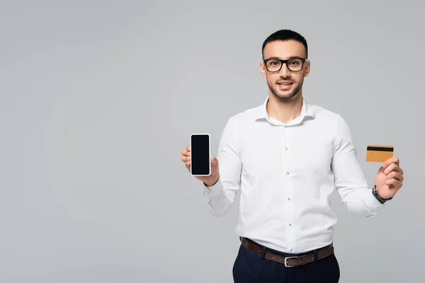 Cheerful hispanic businessman holding credit card and smartphone with blank screen isolated on grey — Stock Photo