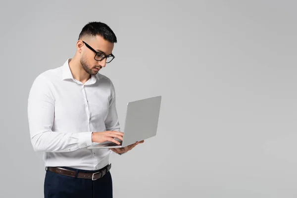 Giovane manager ispanico in occhiali da vista digitando sul computer portatile isolato su grigio — Foto stock