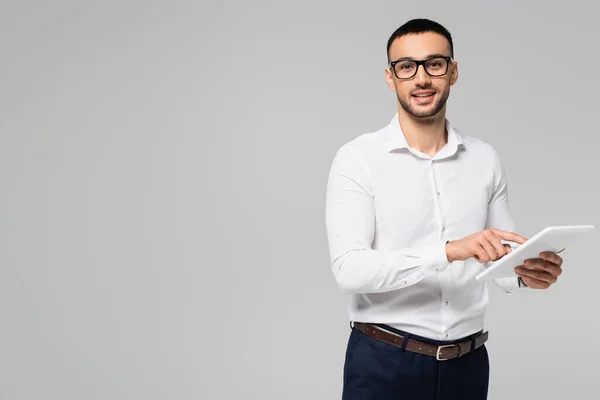 Улыбающийся латиноамериканец в белой рубашке и очках с помощью цифрового планшета, изолированного на сером — стоковое фото