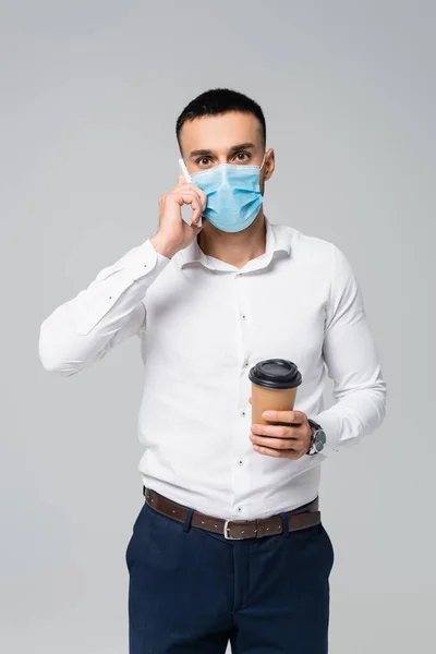 Junger hispanischer Geschäftsmann in medizinischer Maske spricht auf Smartphone und hält Kaffee to go auf grau — Stockfoto